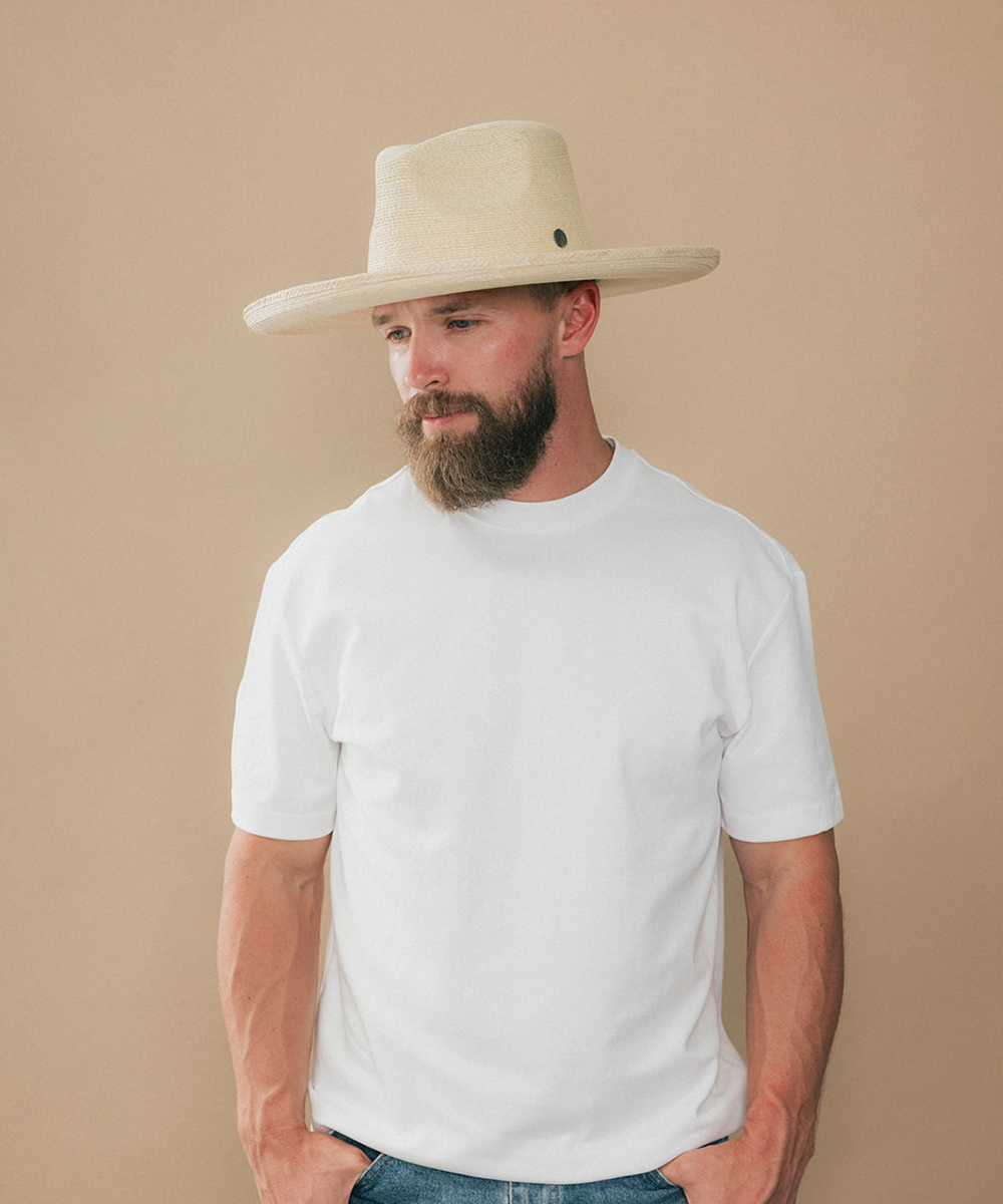 A man with a beard wearing a white crewneck t-shirt and a light beige cowboy hat, standing against a solid beige background #color_ivory