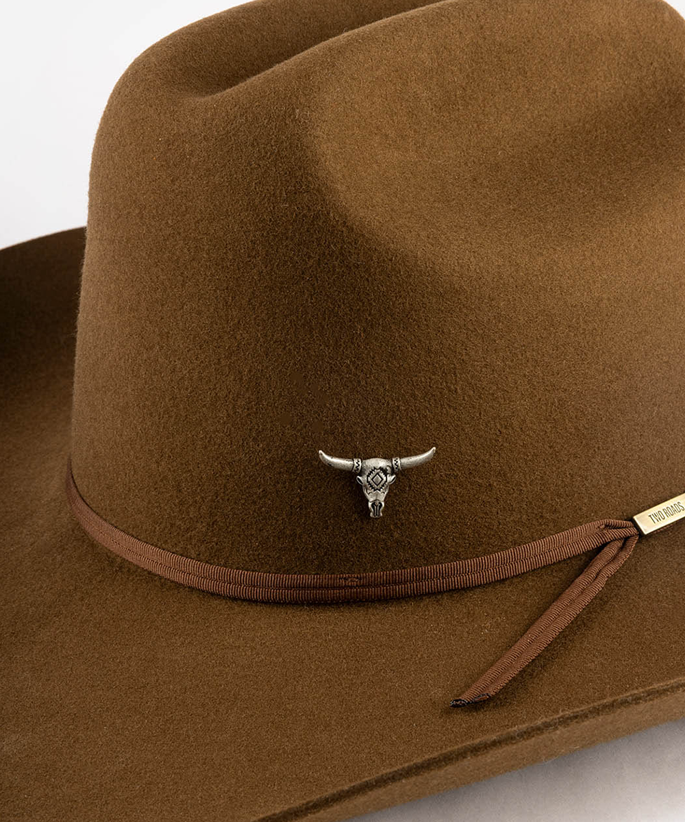 A close-up of a brown felt cowboy hat featuring a brown ribbon band and a silver bull skull pin on the front #color_anti silver