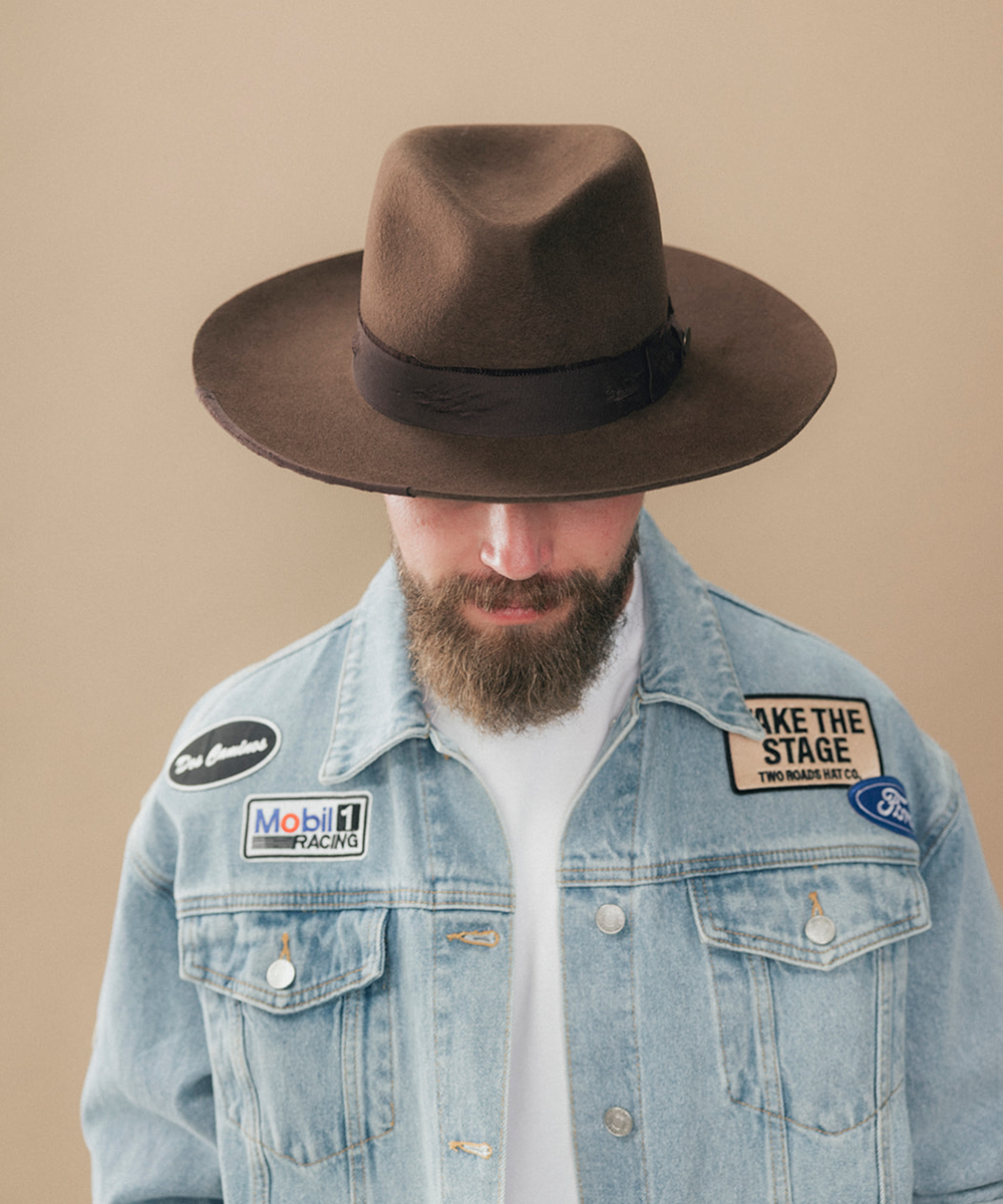 A man with a full beard wearing a dark brown felt fedora hat and a denim jacket and a white t-shirt, shown in a side profile against a solid beige background #color_dark brown