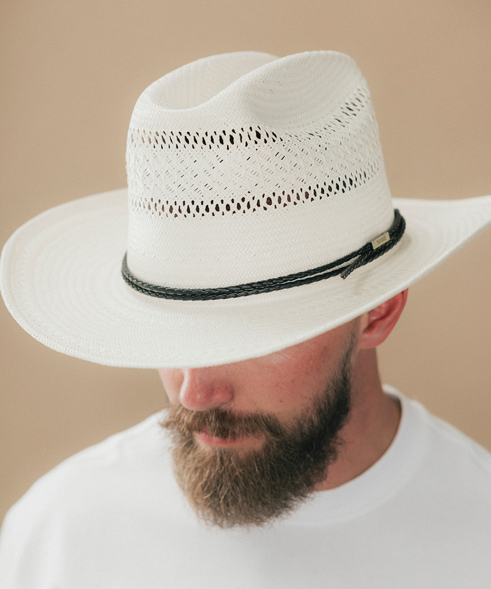A close-up of a man wearing a white woven cowboy hat with a black braided band, against a warm beige background #color_ivory