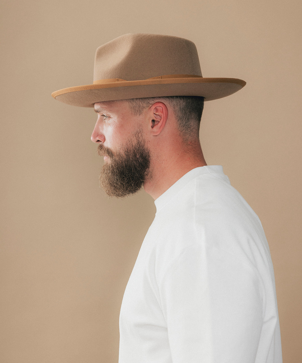 A man in profile wearing a tan wide-brim fedora hat and a white shirt, photographed against a plain beige studio background #color_brown