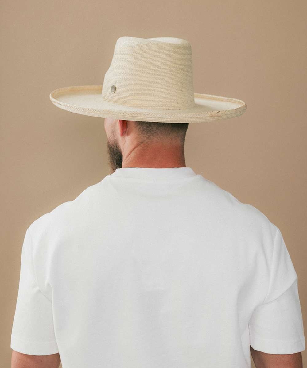 A man viewed from behind wearing a light beige straw cowboy hat and a white t-shirt, set against a solid beige background #color_ivory