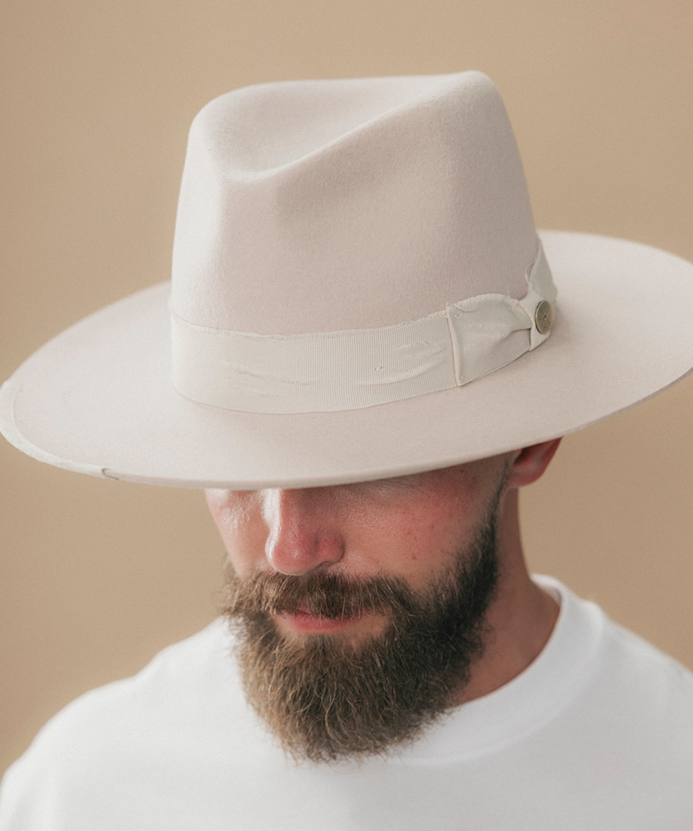 A man with a full beard wearing a cream felt fedora hat and a white t-shirt, shown in a side profile against a solid beige background #color_ivory