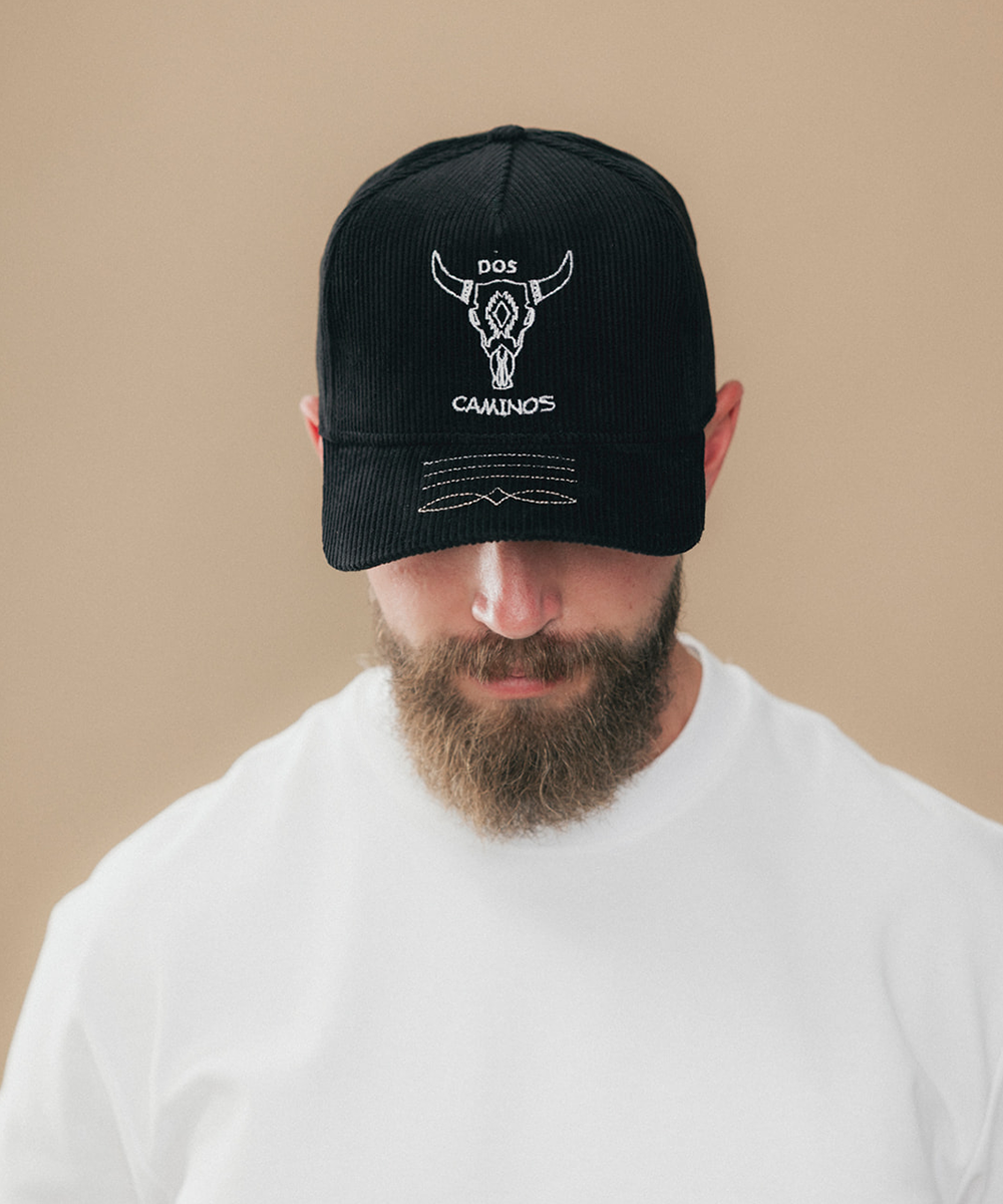 A man wearing a black baseball cap with a white embroidered bull skull logo and the text 'DOS CAMINOS' on the front, paired with a white t-shirt against a solid beige background #color_obsidian