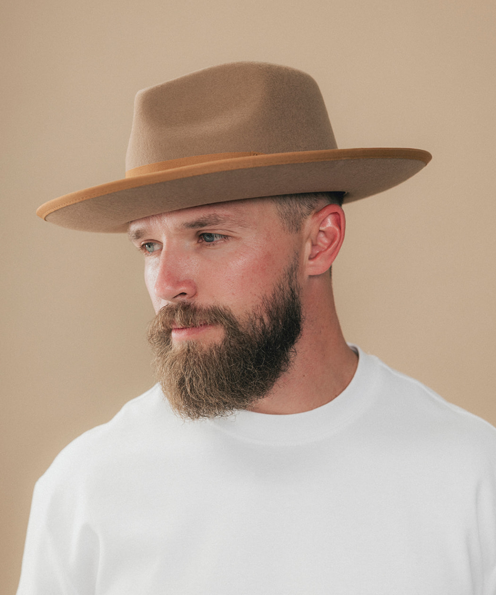 A man with a full beard wearing a light brown wide-brim felt hat and a white t-shirt, shown in a side profile against a solid beige background #color_brown