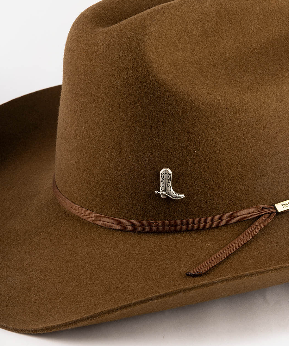 A close-up of a brown felt cowboy hat featuring a matching brown ribbon band with a small silver boot-shaped pin and a gold-tone brand tag #color_anti silver
