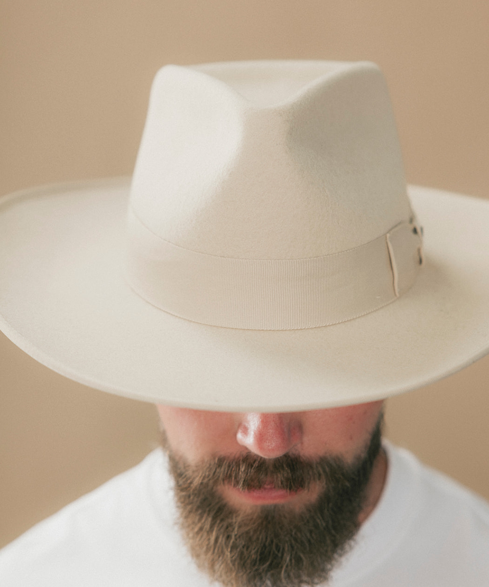 A close-up of a man wearing a wide-brimmed off-white fedora hat, with his beard and lower face visible below the brim against a plain beige background #color_buckskin