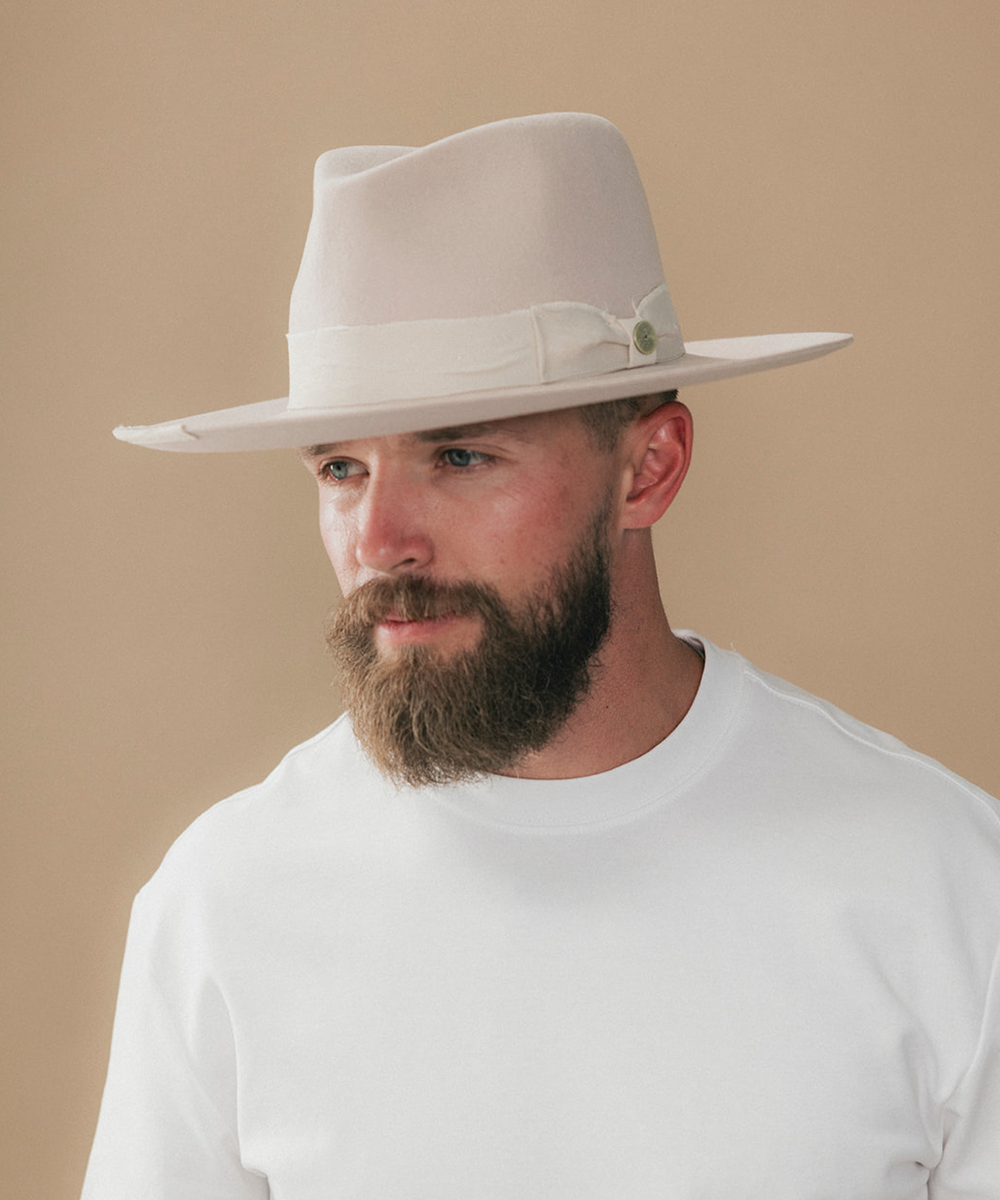 A man with a full beard wearing a cream felt fedora hat and a white t-shirt, shown in a side profile against a solid beige background #color_ivory