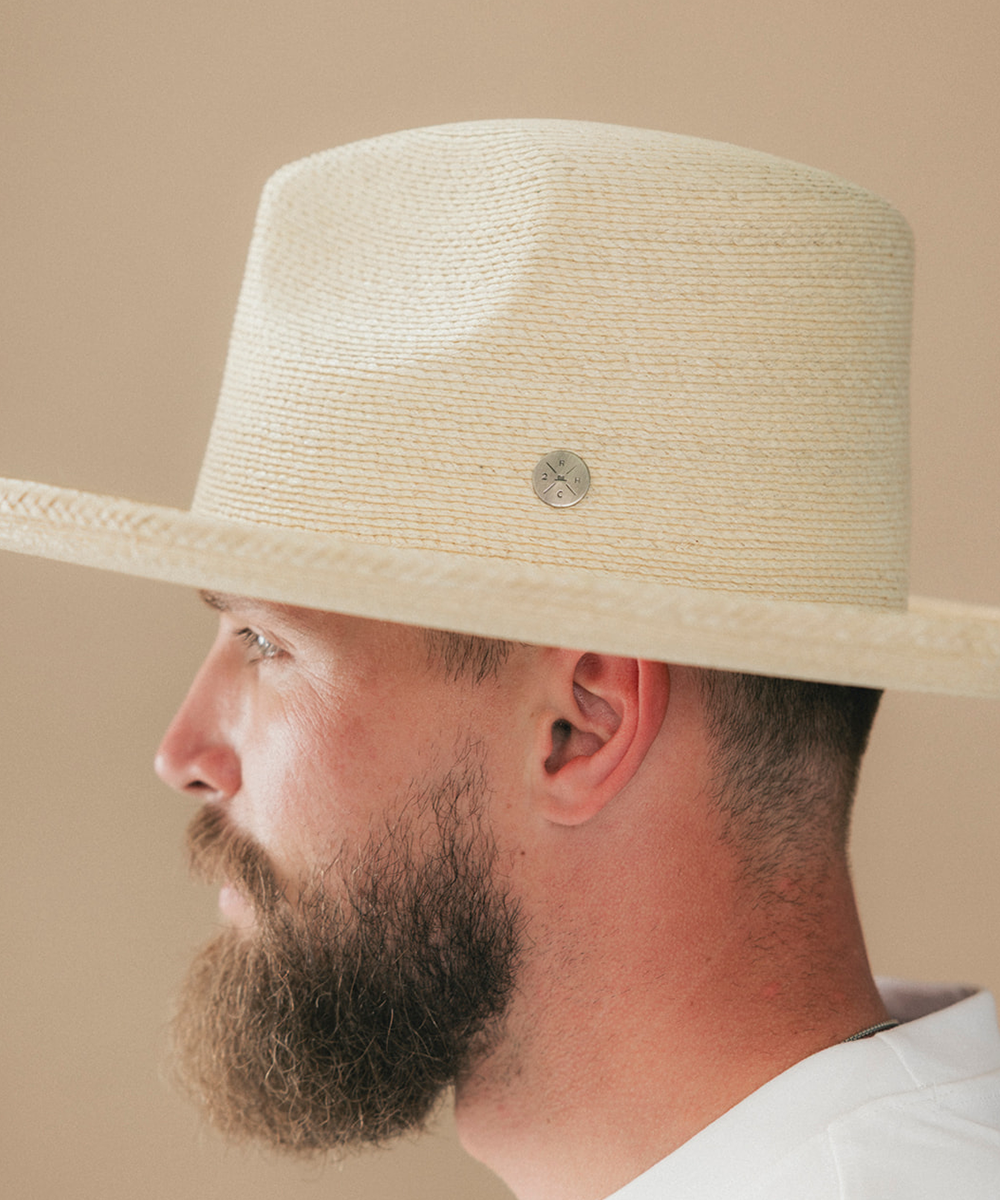 A close-up side profile of a man wearing a light beige woven cowboy hat with a small metallic emblem on the side, set against a solid warm beige background #color_ivory