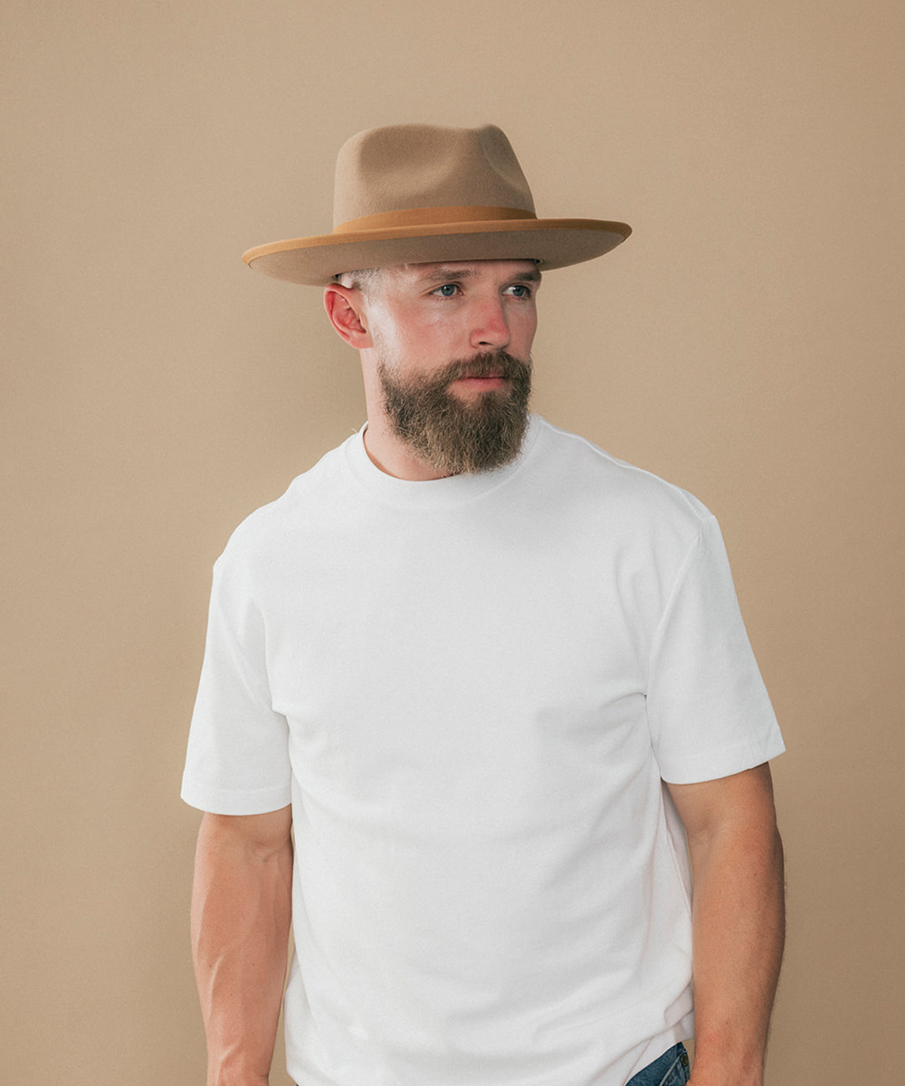 A man with a beard wearing a tan wide-brim fedora hat and a plain white crew-neck t-shirt, standing against a solid beige studio background #color_brown