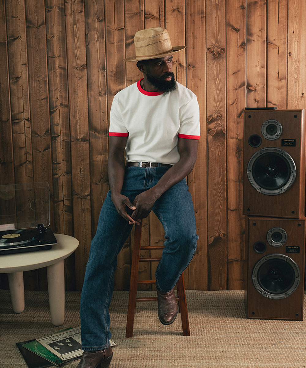 Man sitting on a stool in a room with wooden paneling, wearing a white shirt with red trim and blue jeans #color_natural
