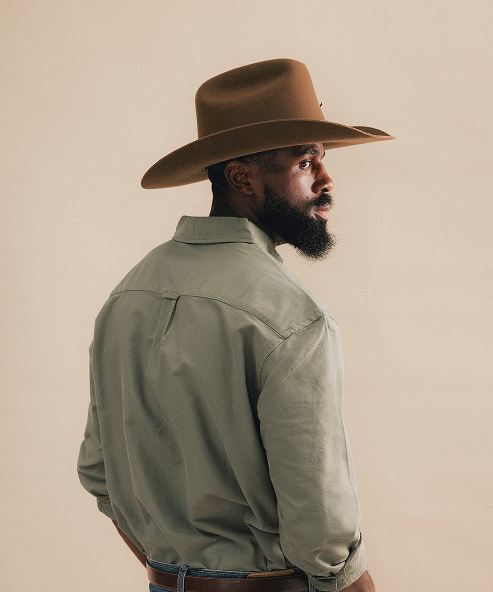 A man wearing a brown wide-brim fedora hat and an olive green long-sleeve button-down shirt, shown from a side-back profile against a solid beige studio background #color_tobacco