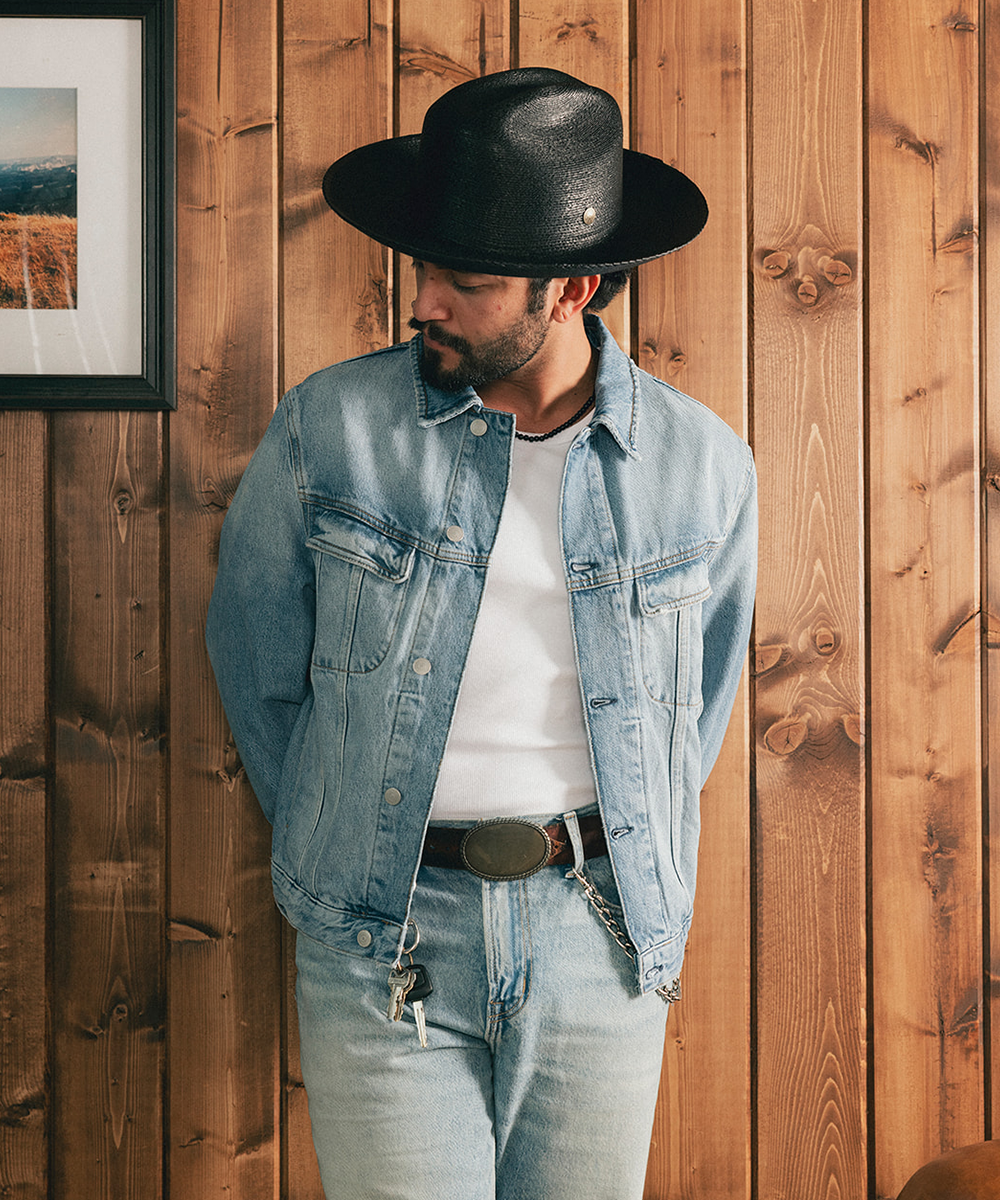 Man wearing a black straw hat and denim jacket against a wooden wall #color_black