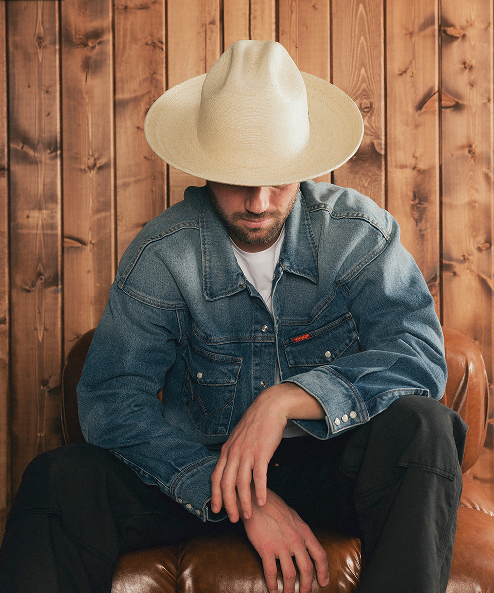 Man wearing a denim jacket and an ivory straw hat sitting on a leather chair against a wooden paneled wall #color_ivory