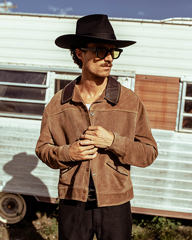 Man wearing a brown suede jacket and black hat standing in front of a vintage trailer.