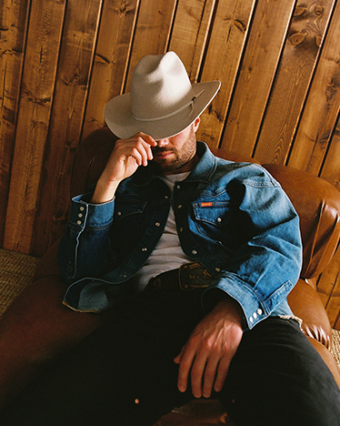 Man wearing a denim jacket and cowboy hat sitting against a wooden wall.