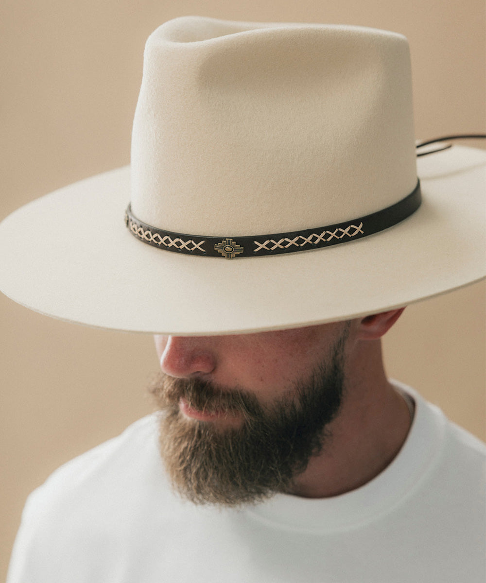 A close-up of a man wearing a wide-brimmed cream-colored felt cowboy hat with a black band decorated with gold-toned geometric patterns and a central star emblem #color_dark brown
