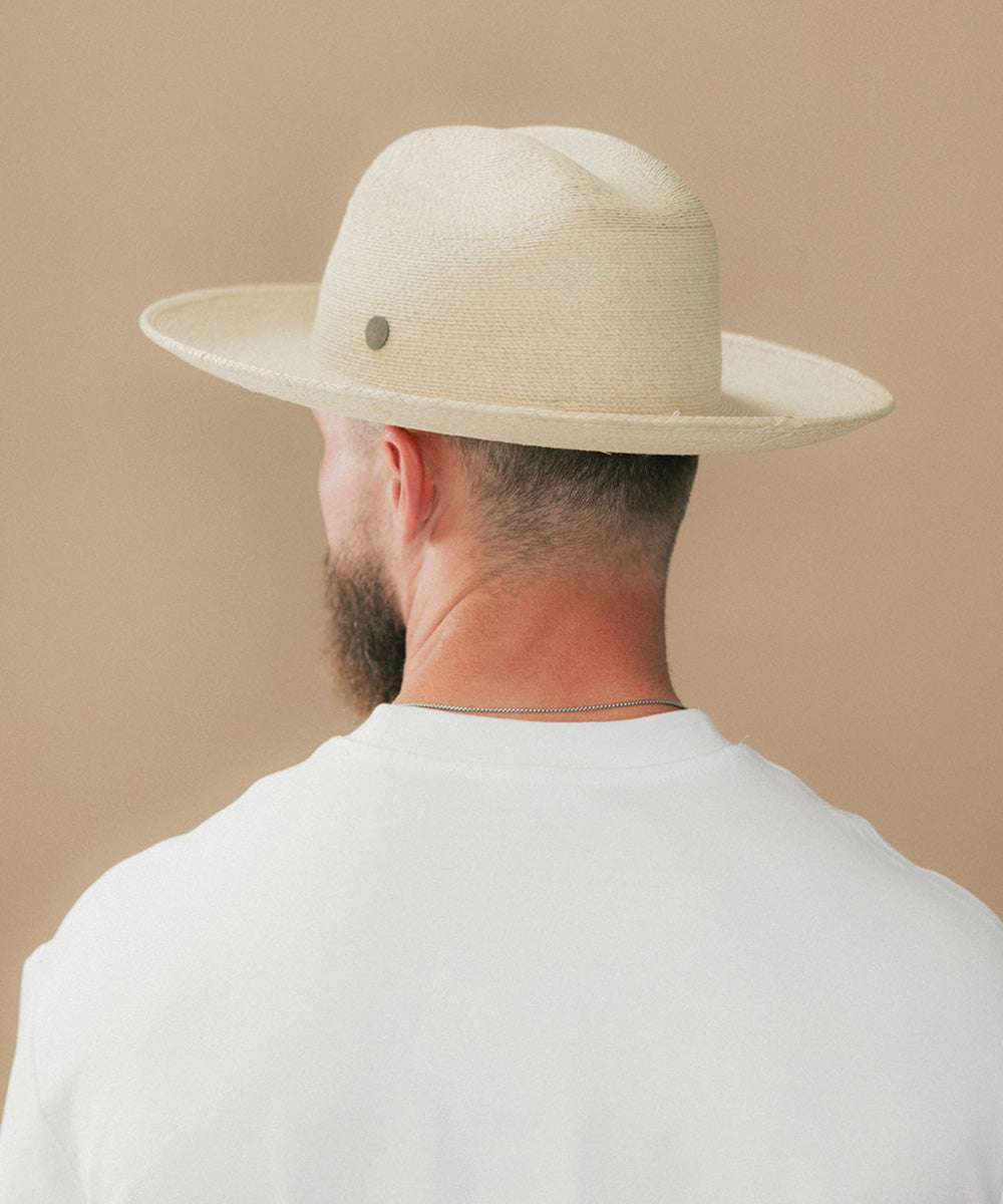 A man viewed from behind wearing a light beige straw fedora hat with a small dark button and a white t-shirt, set against a solid beige background #color_ivory