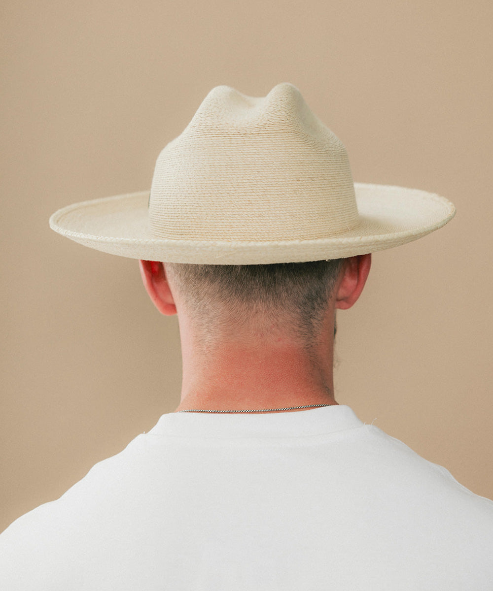 A man viewed from behind wearing a light beige woven cowboy hat and a white t-shirt against a solid beige background #color_ivory
