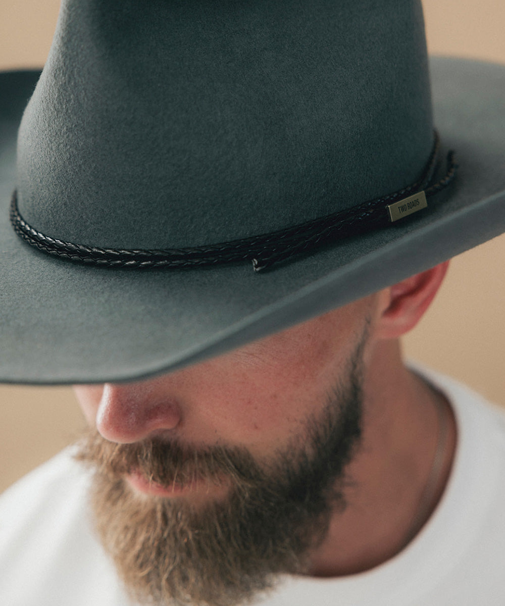 Close-up of a man wearing a dark gray felt cowboy hat with a black braided band and a small metal brand tag #color_black