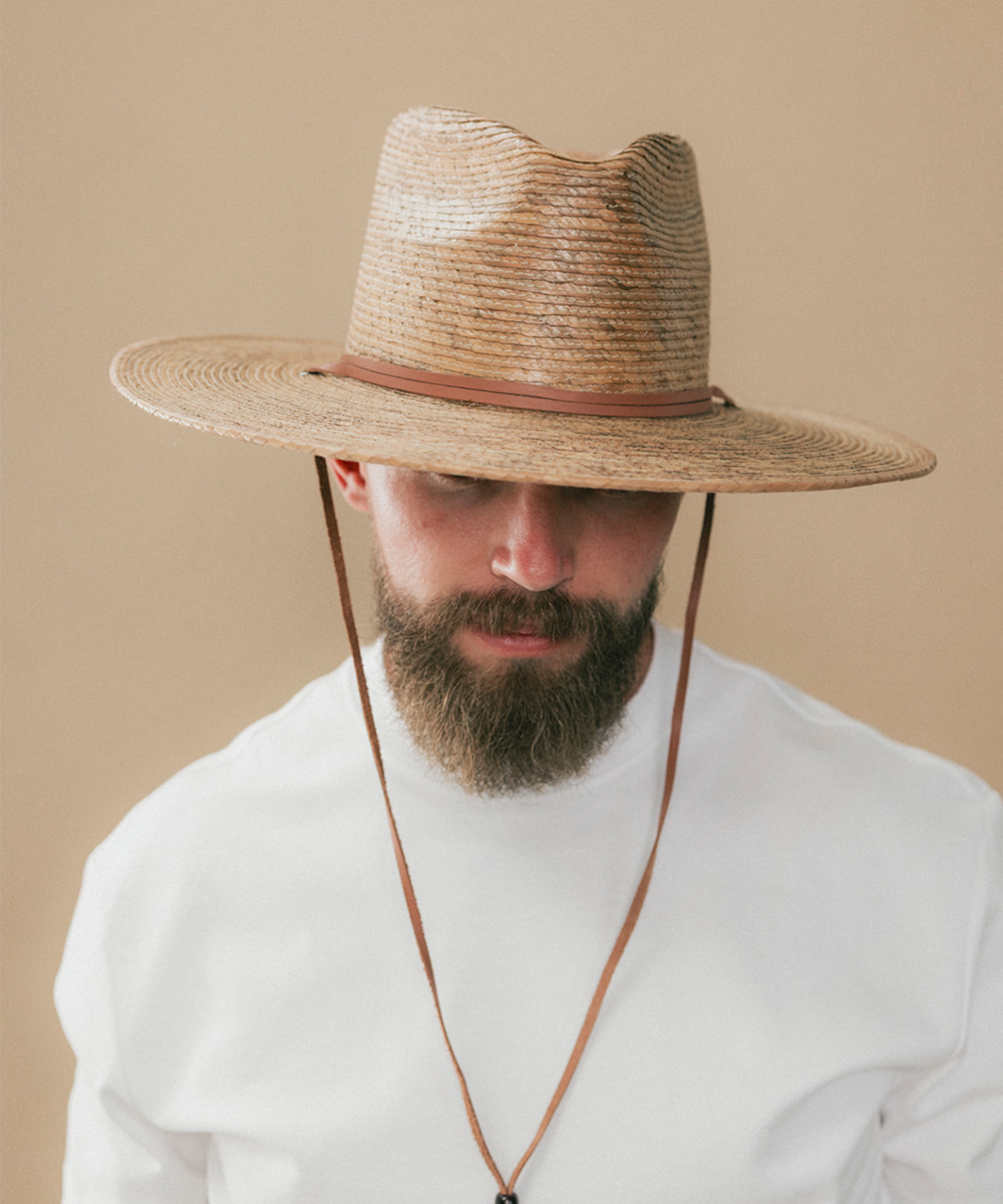 A man wearing a wide-brimmed straw cowboy hat with a brown leather chin strap, paired with a white shirt against a solid beige background #color_toasted