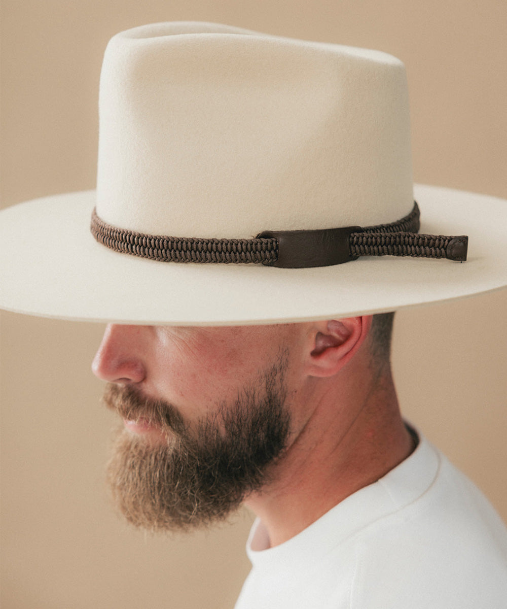 A close-up side profile of a man wearing a wide-brimmed off-white fedora hat with a dark brown woven band and a dark brown bow tie, set against a solid beige background #color_dark brown