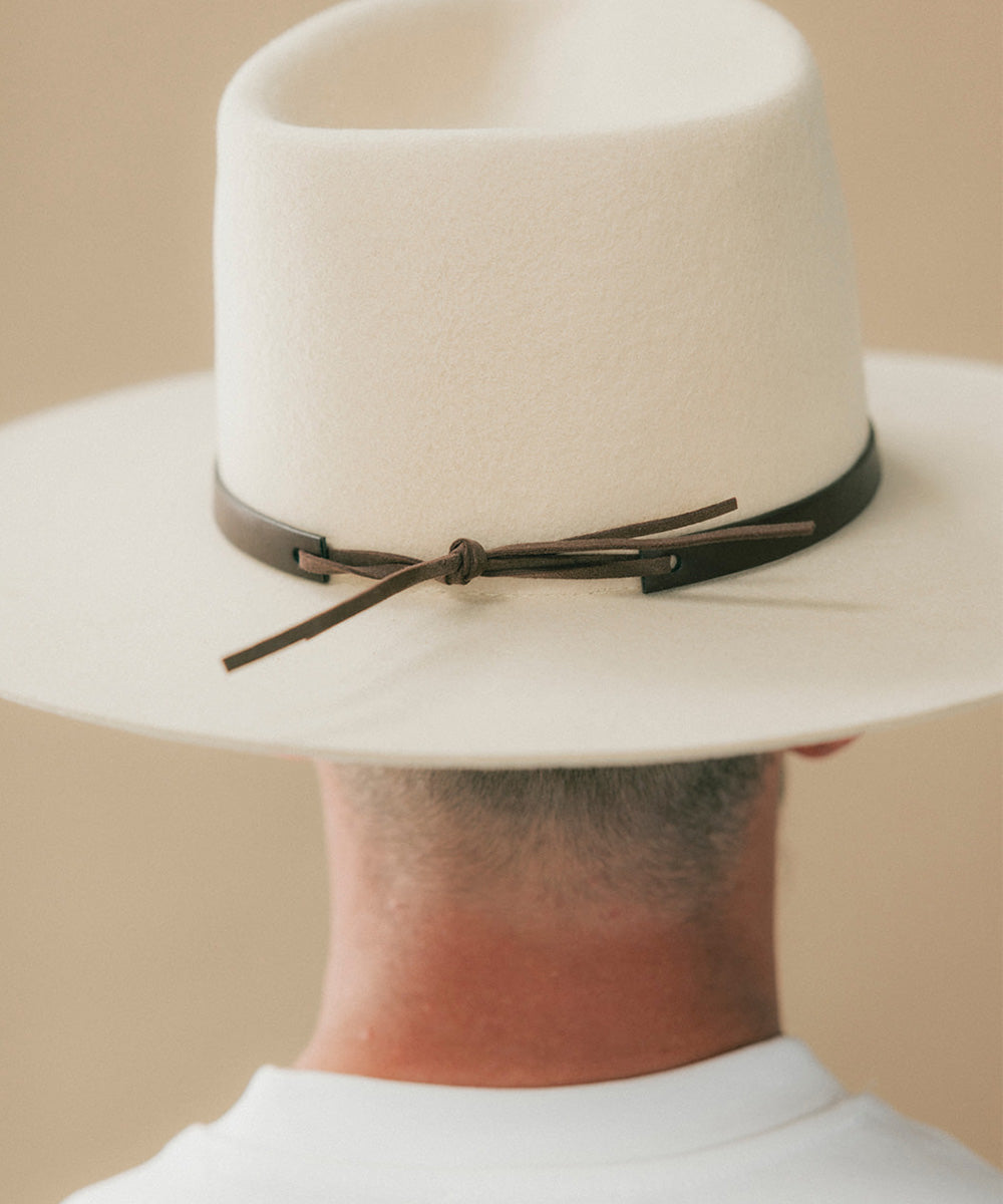 A close-up back view of a person wearing a wide-brim fedora hat in off-white felt with a dark brown leather band tied in a bow around the crown #color_dark brown