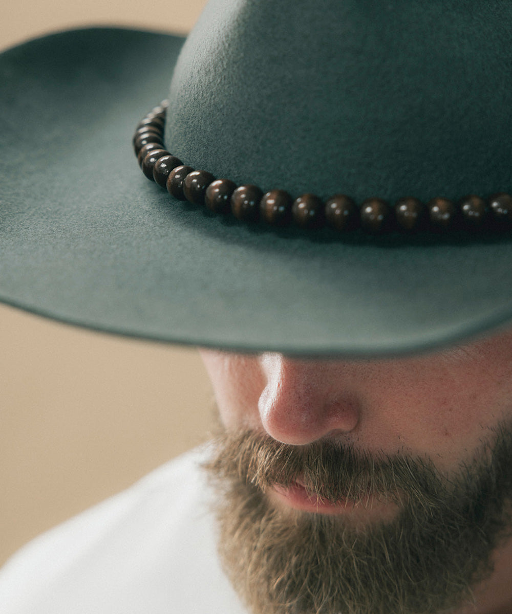 Close-up of a man wearing a dark teal felt hat with a dark brown beaded band, with his beard and nose visible below the brim #color_brown