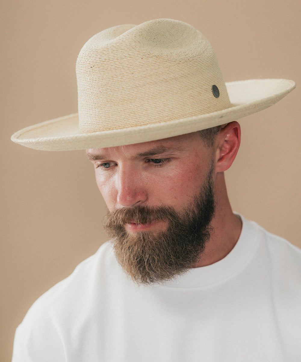 A man with a full beard wearing a light beige woven cowboy hat and a white t-shirt, looking down against a solid beige background #color_ivory