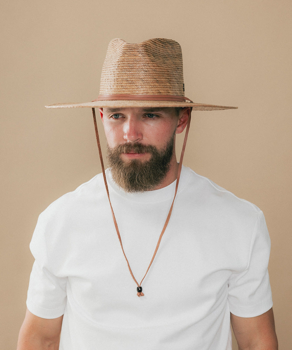 A man wearing a natural straw cowboy hat with a brown leather chin strap, a white t-shirt, and a long brown cord necklace with a small dark bead pendant, set against a solid beige background #color_toasted