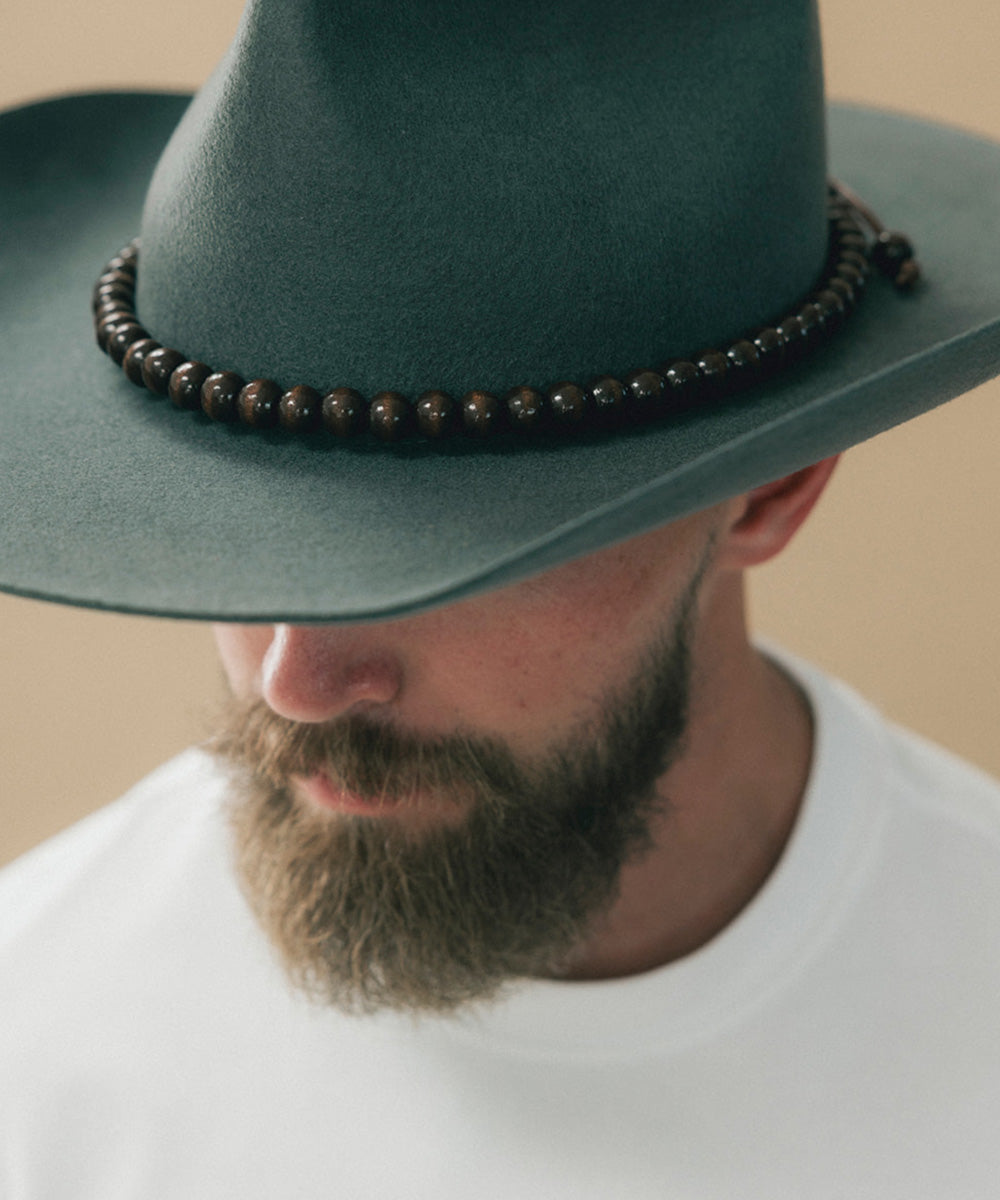 A close-up of a man wearing a dark green wide-brim hat with a dark brown beaded band and a white t-shirt against a plain beige background #color_brown