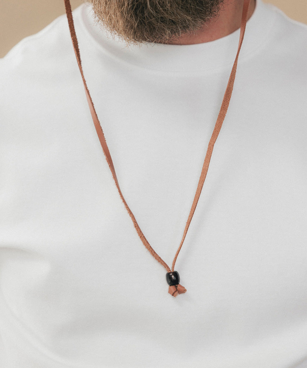 A close-up of a person wearing a thin brown cord necklace with a small black bead and a small brown tassel, over a white t-shirt #color_toasted