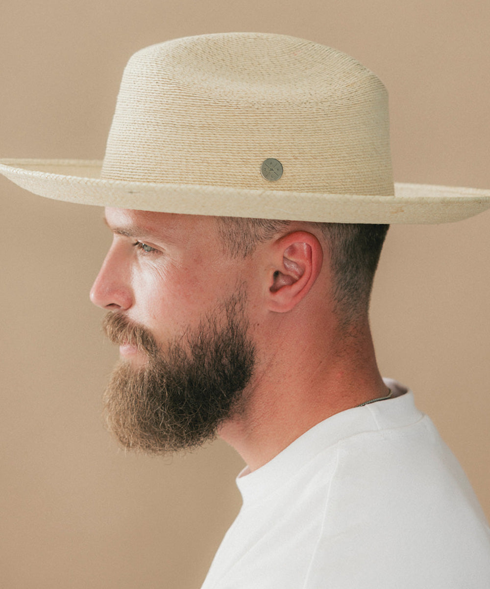 A man in profile wearing a light beige woven cowboy hat with a small round metal button and a white t-shirt, set against a solid beige background #color_ivory