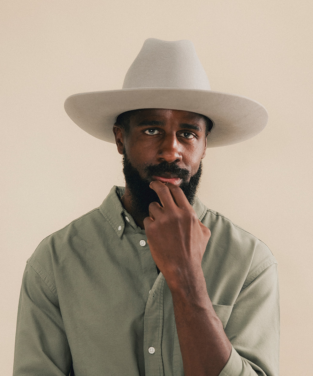 A man with a beard wearing a light gray wide-brim hat and an olive green button-down shirt, posing against a solid beige background #color_stone