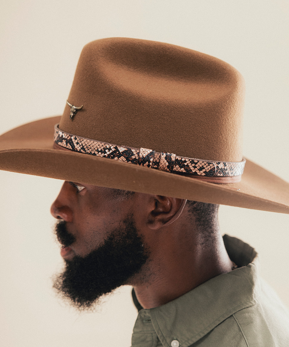 A man in profile wearing a brown felt cowboy hat with a patterned band and a small silver bull skull pin, against a plain light beige background #color_brown