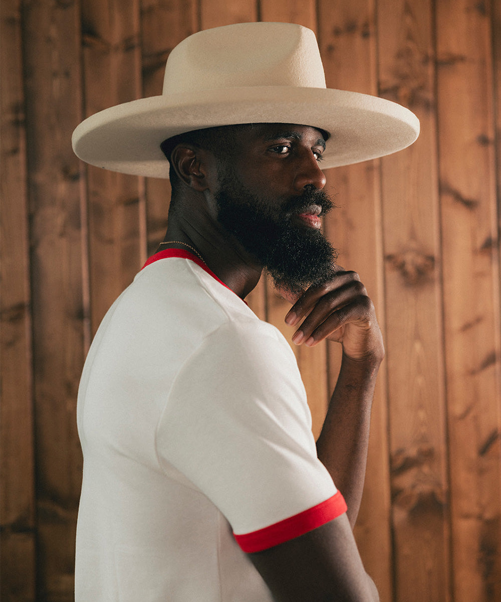 Man wearing a white hat and white shirt with red trim against a wooden background #color_buckskin