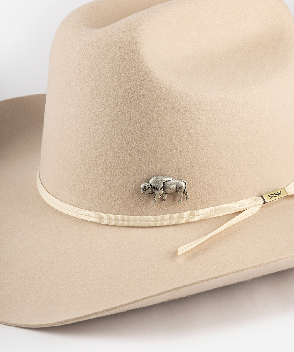 A close-up of a beige felt cowboy hat featuring a matching beige ribbon band with a small silver buffalo pin and a gold-tone brand tag #color_anti silver
