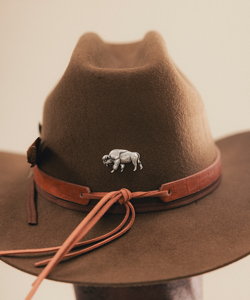 A brown felt cowboy hat with a brown leather band and tie, featuring a small silver metal bison pin on the side, photographed against a plain light beige background #color_anti silver