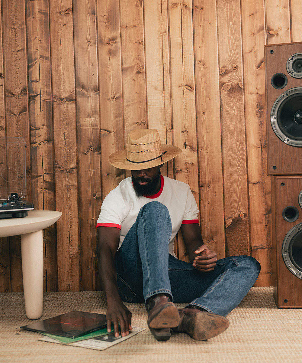 Man sitting on the floor with a straw hat, surrounded by vintage speakers and records against a wooden wall #color_natural