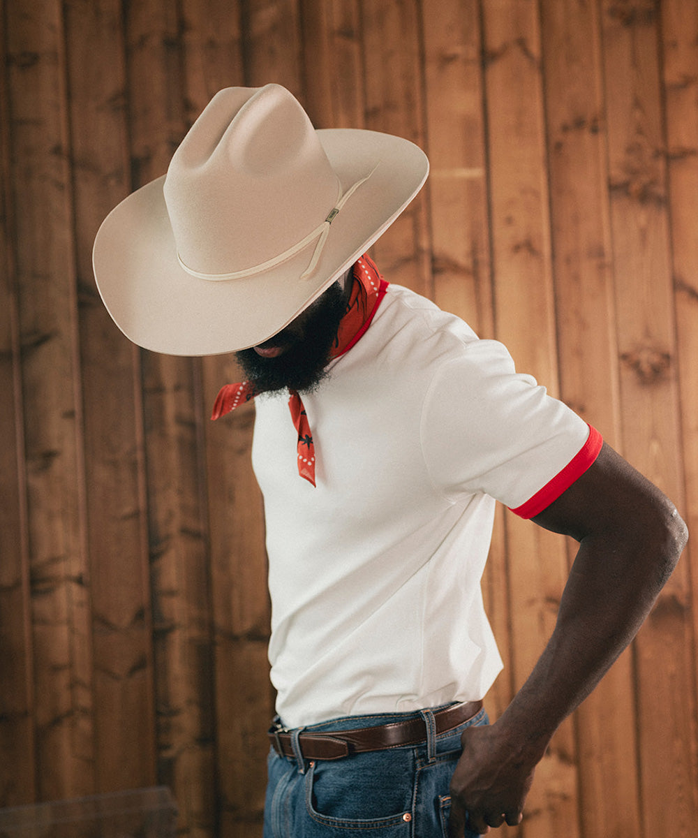 Man wearing a cowboy hat and white shirt with red trim against a wooden background #color_buckskin