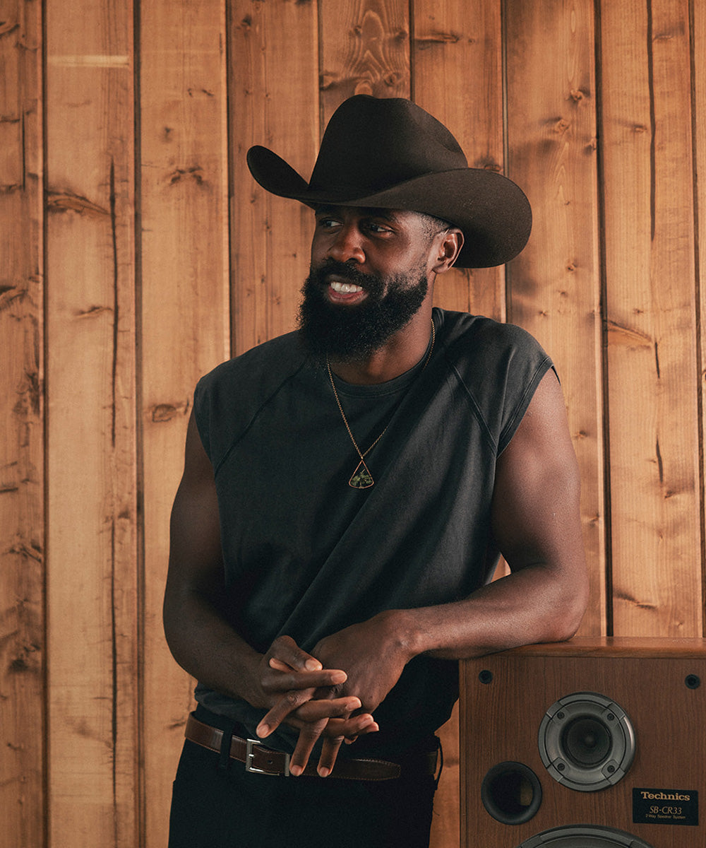 Man wearing a black cowboy hat and sleeveless shirt standing against a wooden paneled wall #color_hickory