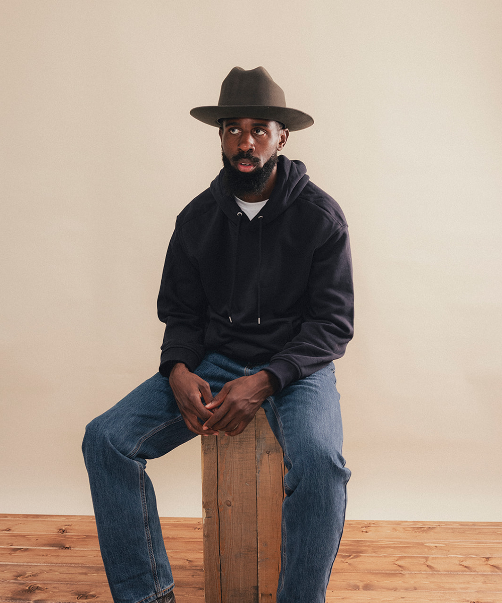 A man wearing a dark navy pullover hoodie, blue jeans, and a brown wide-brim hat sits on a wooden crate against a plain beige wall and wood floor #color_hickory