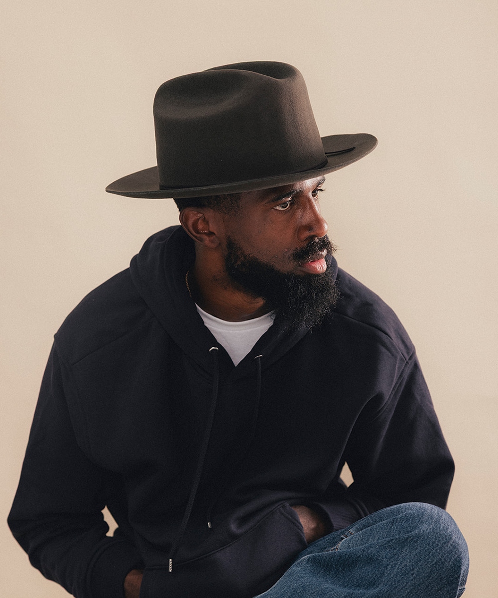 A man wearing a dark brown wide-brim fedora hat, a black pullover hoodie over a white t-shirt, and blue jeans, posed against a solid beige background #color_hickory