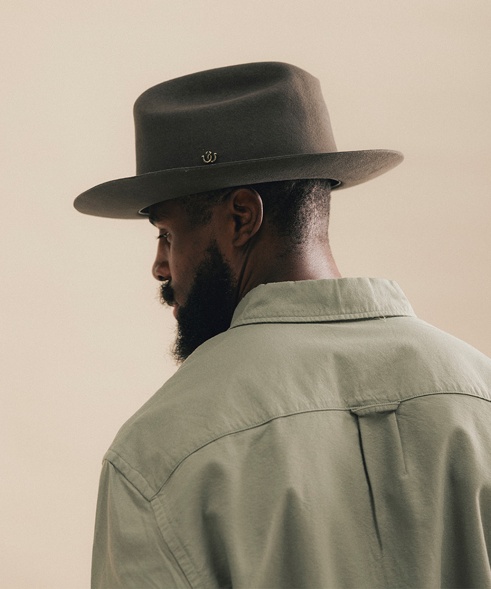 A man in profile wearing a dark brown felt fedora hat with a small gold logo and a light sage green button-up shirt, set against a solid warm beige background #color_hickory
