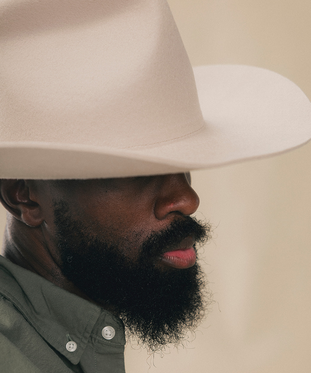 A man wearing a wide-brimmed cream cowboy hat and an olive green button-down shirt, photographed against a solid beige background #color_silverbelly