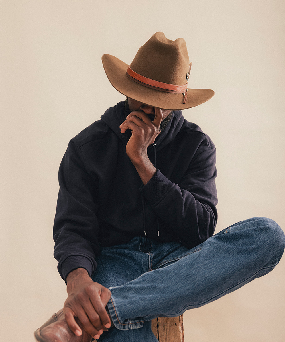 A man wearing a brown felt cowboy hat with a pink band, a black hoodie, and blue jeans sits cross-legged against a plain beige background #color_tobacco