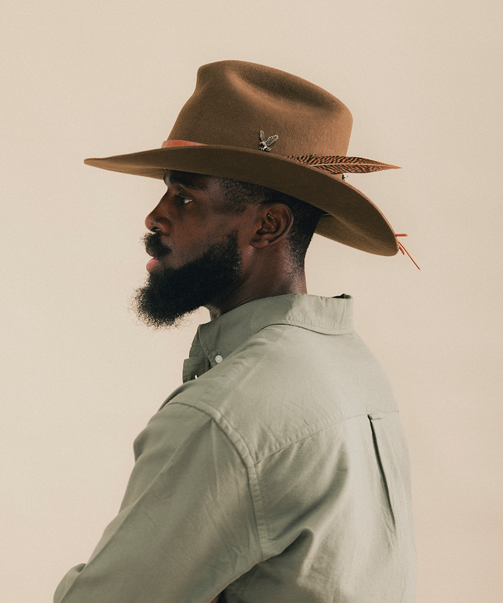A man in profile wearing a brown felt cowboy hat with a feather accent and a light sage green button-up shirt, set against a solid beige background #color_tobacco