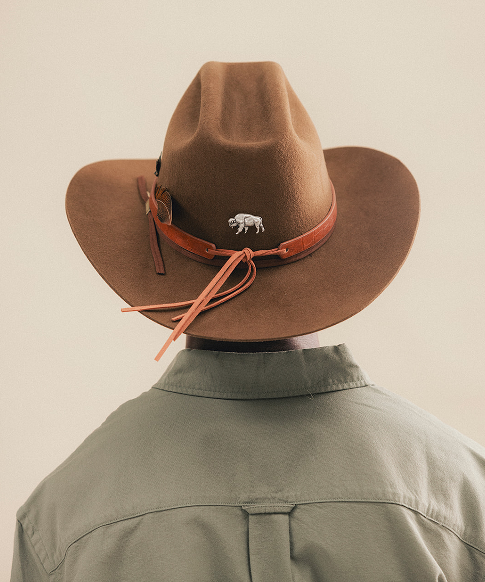 A brown felt cowboy hat with a tan leather band and tie, decorated with a small white buffalo pin, shown on a model from the back wearing an olive green shirt against a light beige background #color_tobacco