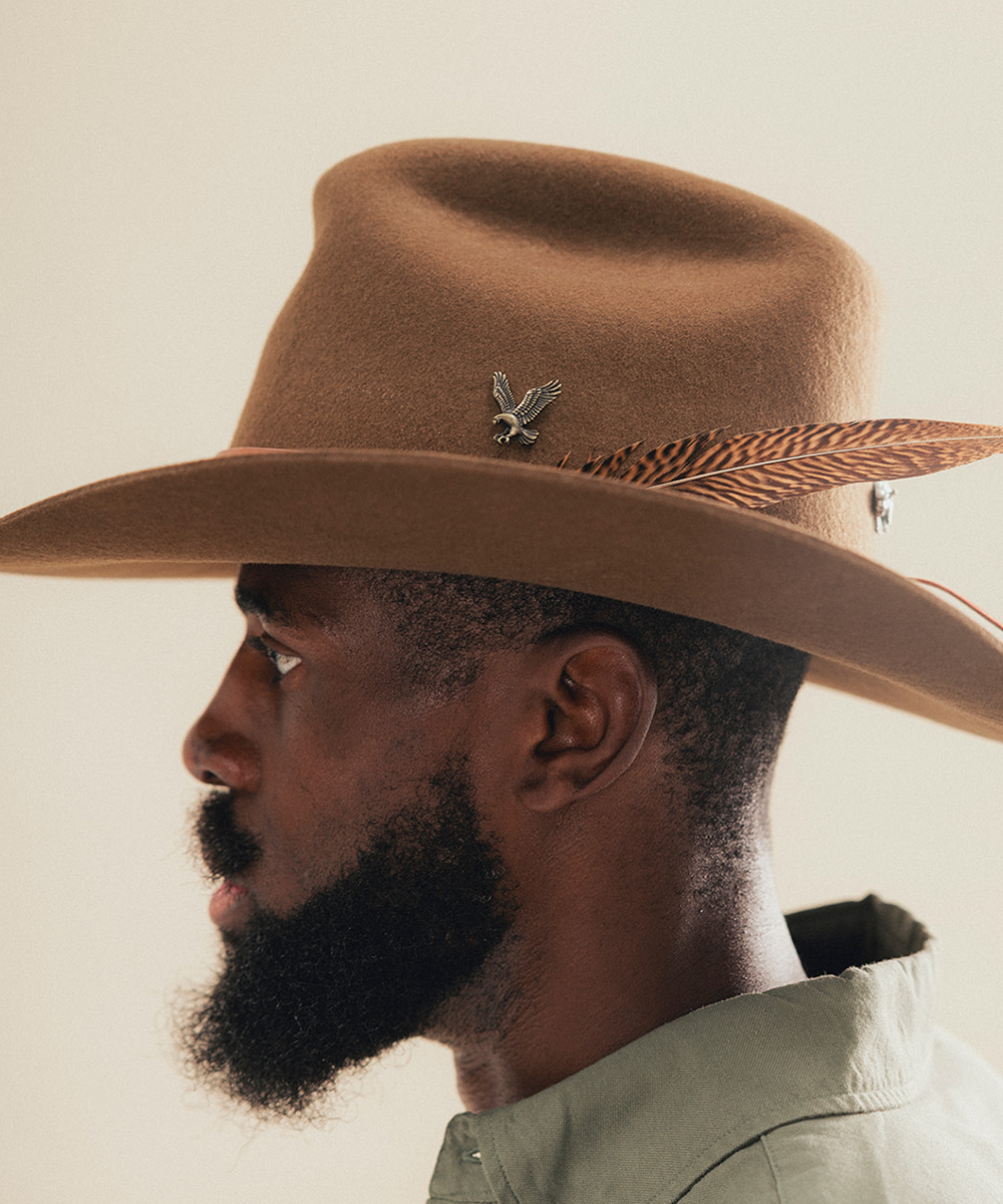 A man in profile wearing a brown felt cowboy hat with a feather and small metallic bird accent, against a plain light beige background #color_tobacco