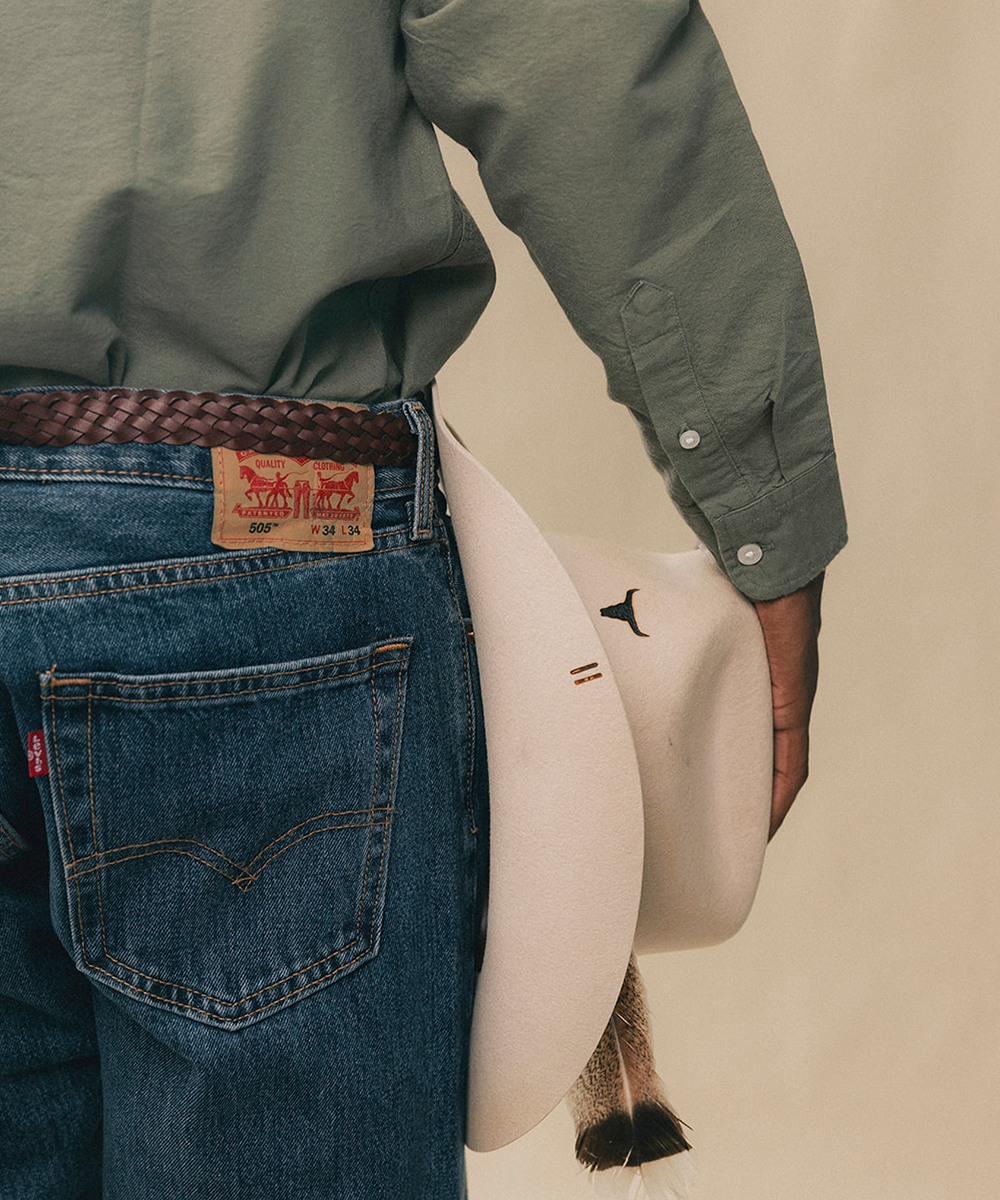 Close-up of a person wearing blue Levi's jeans, an olive green shirt, a brown braided belt, and holding a cream-colored cowboy hat against a warm beige background #color_buckskin